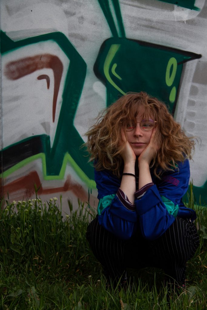 Szabó Boglárka fiatal képzőművész szemüvegben, kék kabátban, zöld és szürke graffiti előtt